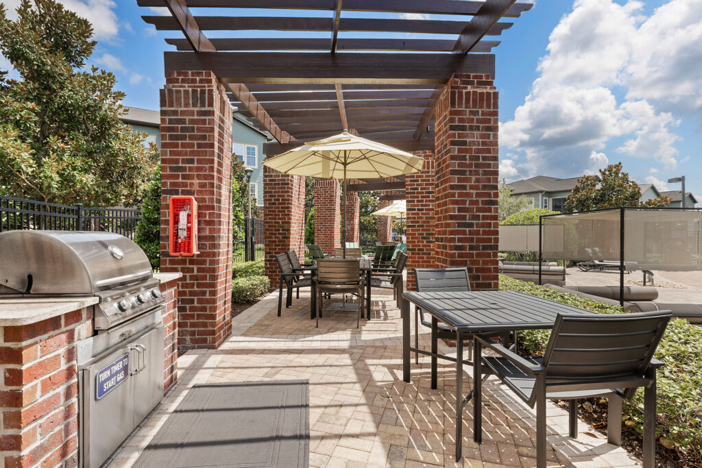 Outdoor patio with brick pillars, a grill, dining tables, chairs, and a large umbrella. The area is sunny, surrounded by greenery, and includes a fire extinguisher mounted on a pillar.