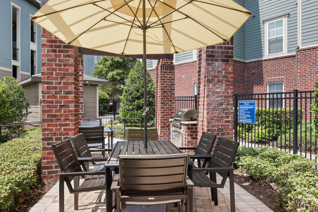 Outdoor patio area with a black table, eight chairs, and a large beige umbrella. Brick columns, a barbecue grill, greenery, and apartment buildings surround the space. A blue sign is visible on the fence in the background.