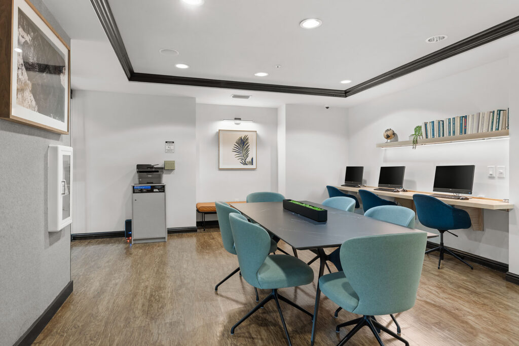 Modern office space with a black table surrounded by teal chairs, three computers on a wooden desk against the wall, a printer in the corner, and minimal décor including framed art and a shelf with books.