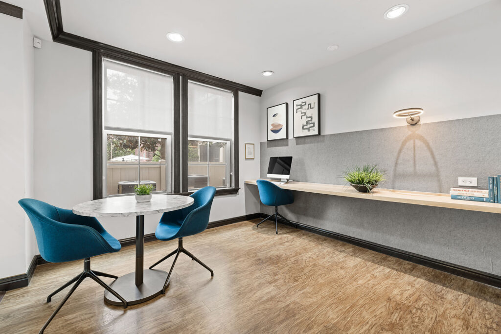 Modern office with wood floor, two blue chairs at a round marble table, built-in desk with books, plant, iMac, and wall art. Large windows let in natural light; neutral walls and ceiling lights brighten the space.