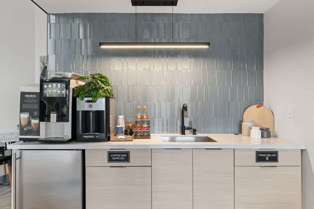 Modern coffee bar with a Keurig machine, syrup bottles, cups, a sink, and a plant on a light wood counter against a gray tiled backsplash. Cabinets below are labeled for coffee supplies and trash.