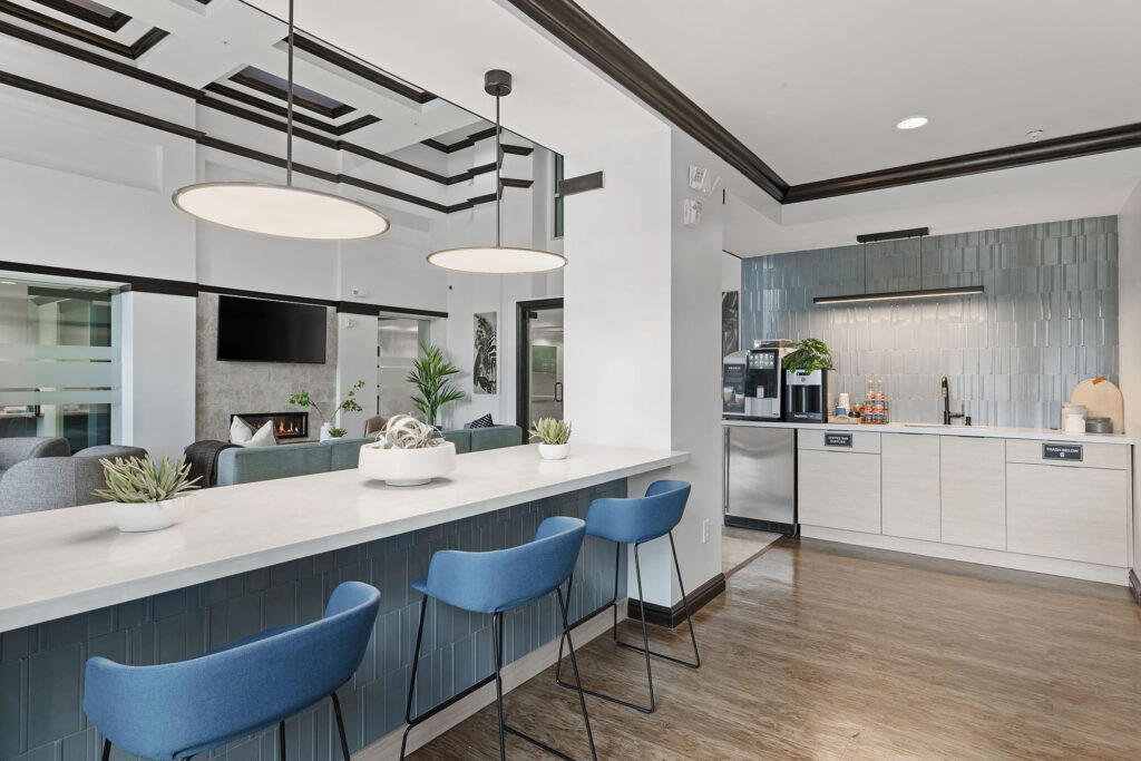 Modern kitchen with blue barstools, a long white countertop, hanging lights, light wood floors, and a coffee station against a blue-tiled backsplash. There are plants as decor and a lounge area visible in the background.