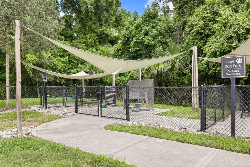 Fenced large dog park with shade sails, benches, signs, and greenery in the background. Signs indicate the area is for dogs over 20 lbs. Concrete walkway leads to the entrance gate.