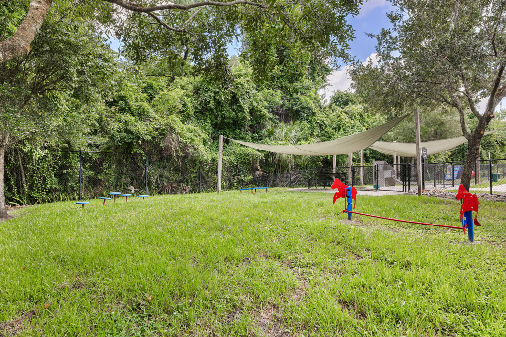 A grassy park area with trees, shaded seating, and playground equipment, including red seesaws. A white canopy provides shade over part of the space. A fence runs along the back.