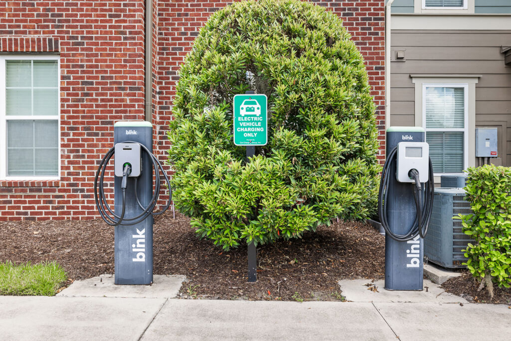 Two Blink electric vehicle charging stations are installed on a sidewalk in front of a large bush. A sign above the bush reads Electric vehicle charging only. Brick and siding exterior walls are visible in the background.