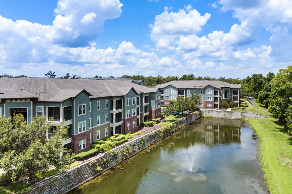 A pond with a water fountain bordered by modern green and brick apartment buildings, lush landscaping, and trees under a blue sky with scattered clouds.