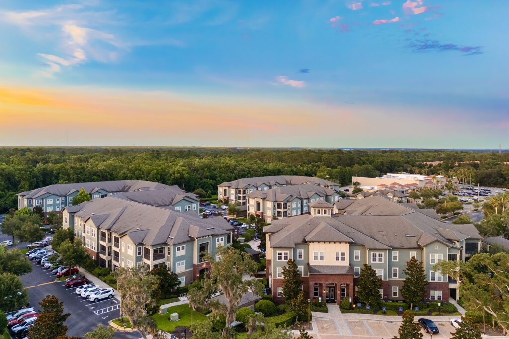 Aerial view of a large apartment complex with multiple buildings, green trees, and parked cars at sunset under a colorful sky.