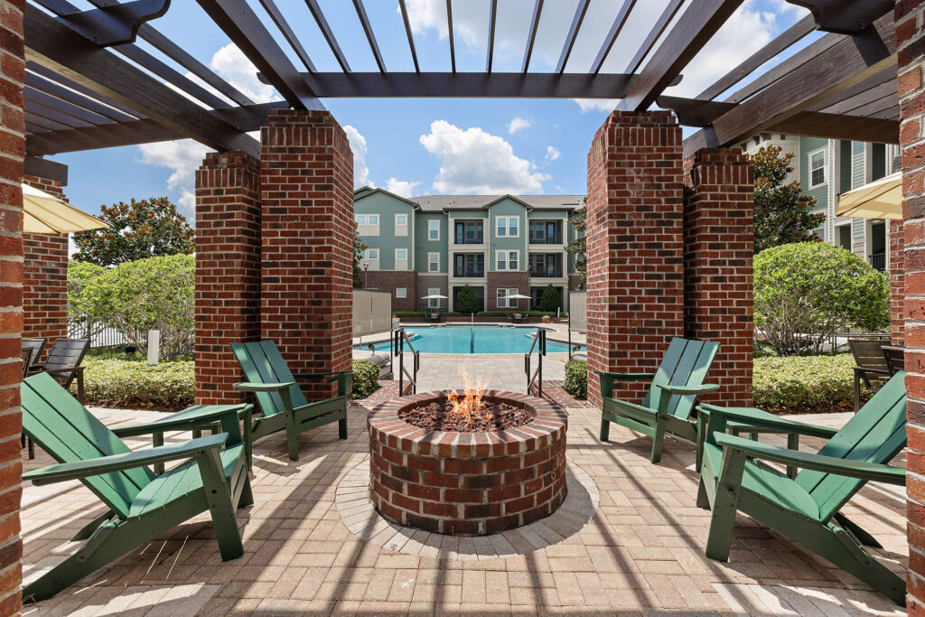 Four green Adirondack chairs surround a brick fire pit under a pergola, with a swimming pool and apartment buildings in the background on a sunny day.