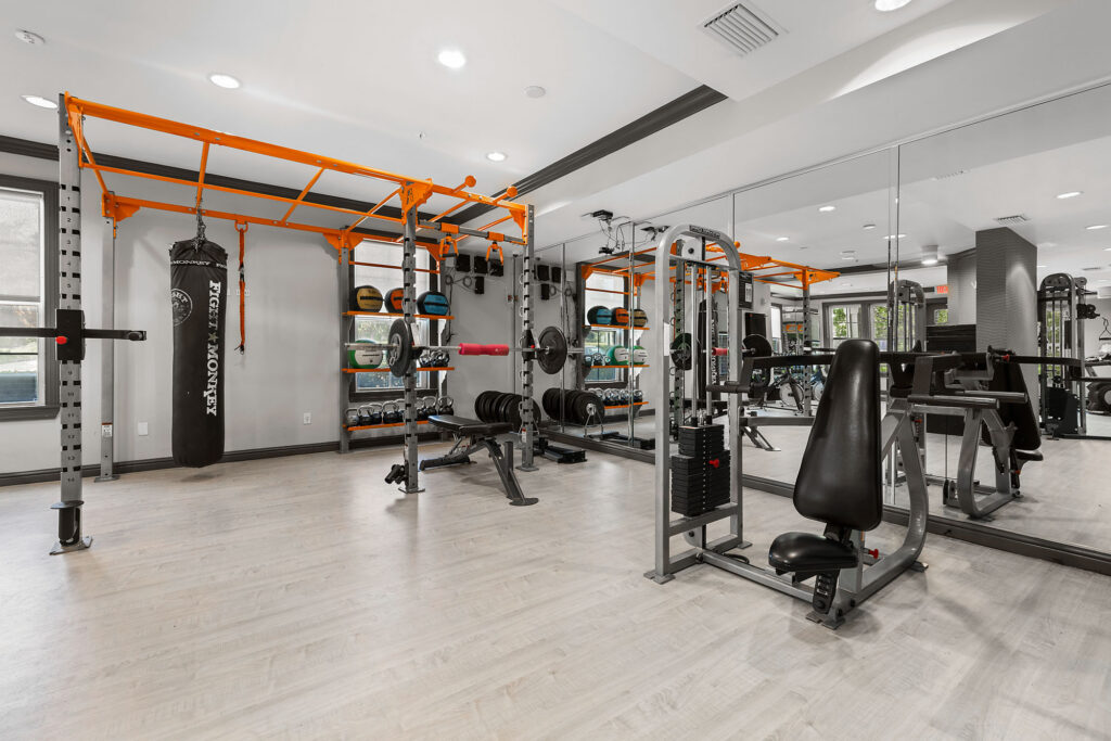 Modern gym with weightlifting equipment, a punching bag, benches, weight plates, cable machines, and large mirrors on the walls. The space has light wood floors and bright lighting.