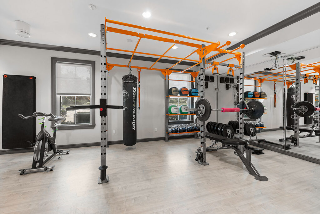 Modern gym with weightlifting rack, punching bag, bench press, exercise bike, free weights, and large mirrors on the wall; bright lighting and light wood flooring.