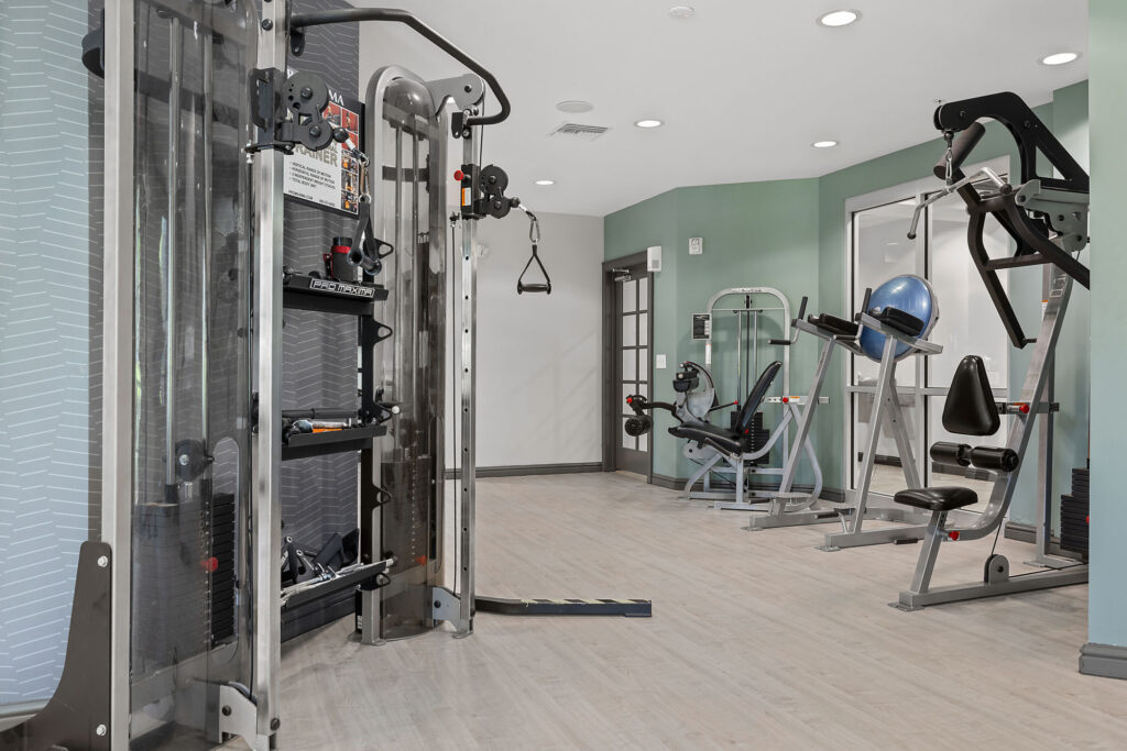 A modern gym with various exercise machines, including a cable machine, seated row, and leg press, on light wood flooring with mint green and white walls.