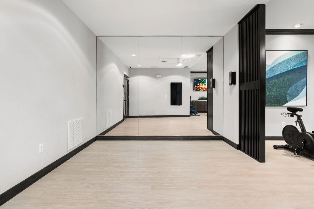 A modern, empty exercise room with mirrors, light wood flooring, white walls, and an exercise bike partially visible on the right side. A colorful abstract artwork hangs on the wall.