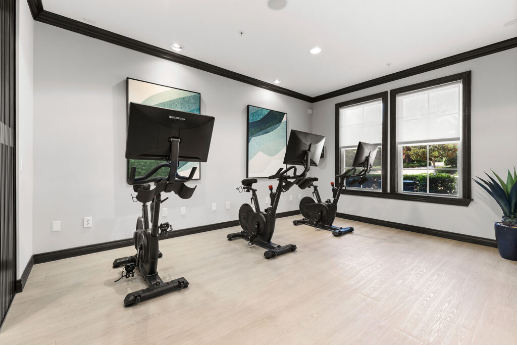 Three stationary exercise bikes with screens are lined up in a bright, modern room with wooden floors, large windows, abstract wall art, and a potted plant in the corner.
