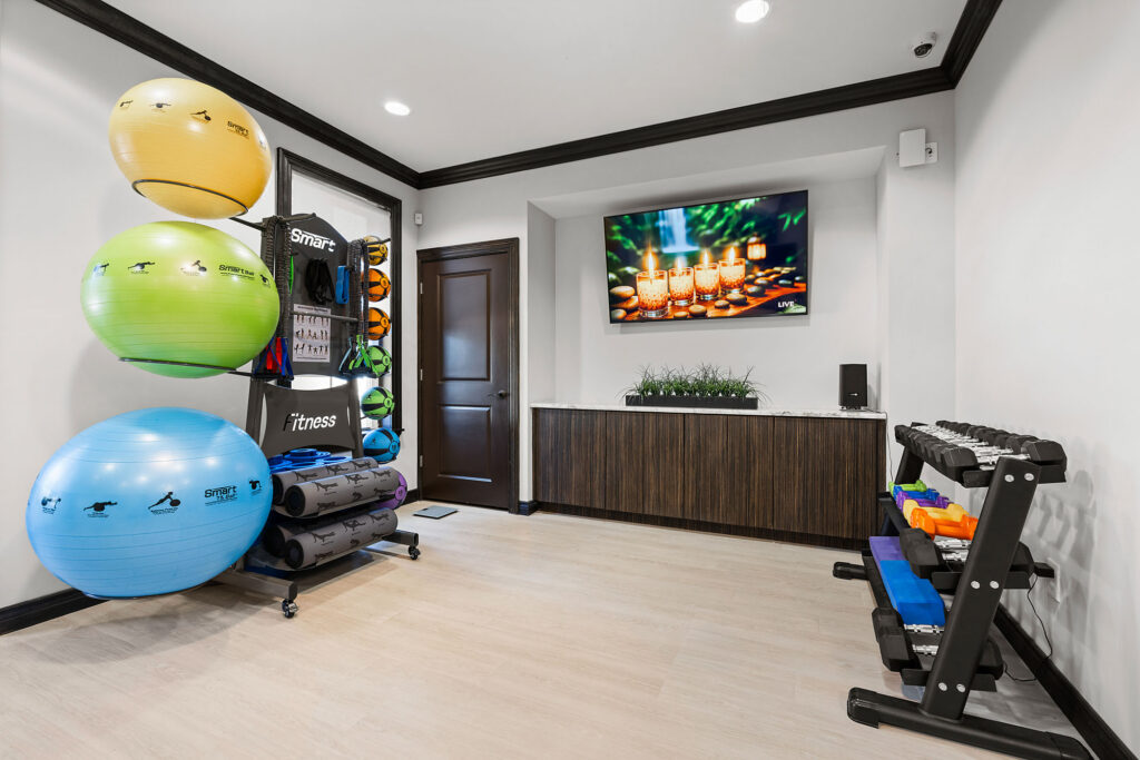 A fitness room with large exercise balls, yoga mats, dumbbells, and a TV displaying candles. The space has light wood floors, white walls, and a dark wooden cabinet with a green plant.