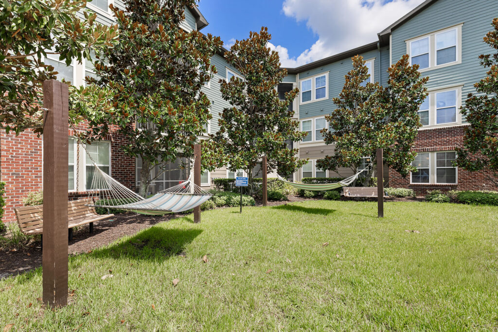 Three hammocks and two benches are set up on a grassy area between trees and a modern apartment building with light blue siding and red brick accents under a partly cloudy sky.