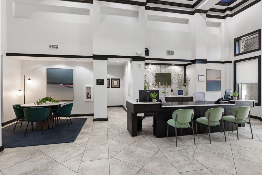 Modern office lobby with a large reception desk, four green chairs, and a marble accent wall. To the left, there’s a round table with four green chairs and two abstract paintings on the walls.