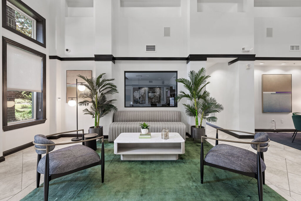 Modern living room with gray chairs, a light sofa, white coffee table, green rug, potted plants, and large windows. Artwork and neutral walls create an inviting, contemporary atmosphere.