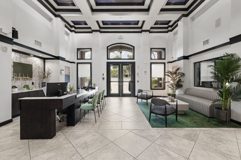 A modern office lobby with a long black reception desk, green chairs, a gray sofa, coffee tables, potted plants, large windows, and white walls with high ceilings and black accents.
