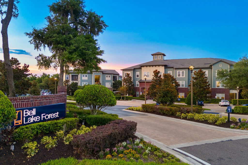 A modern apartment complex at sunset, with manicured landscaping, trees, and a sign reading Bell Lake Forest near the entrance.