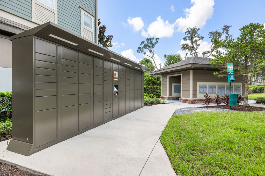 Outdoor package locker system on a sidewalk near an apartment building, with a small utility structure, grass, and trees in the background under a partly cloudy sky.