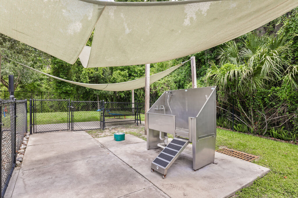 Outdoor pet washing station with a metal tub, stairs, and shade sails, surrounded by a black chain-link fence and lush green trees.
