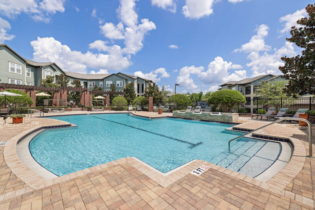 Large outdoor swimming pool with steps, surrounded by lounge chairs, green shrubs, and apartment buildings under a blue sky with scattered clouds. Paved pool deck and safety depth markers are visible.