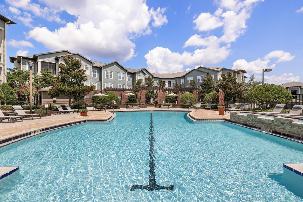 Outdoor swimming pool with clear blue water, surrounded by lounge chairs and landscaped greenery, in front of modern apartment buildings under a partly cloudy sky.