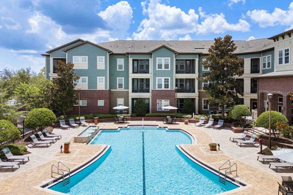 Outdoor swimming pool with lounge chairs and umbrellas, surrounded by landscaped bushes and trees, in front of a multi-story apartment building under a partly cloudy sky.