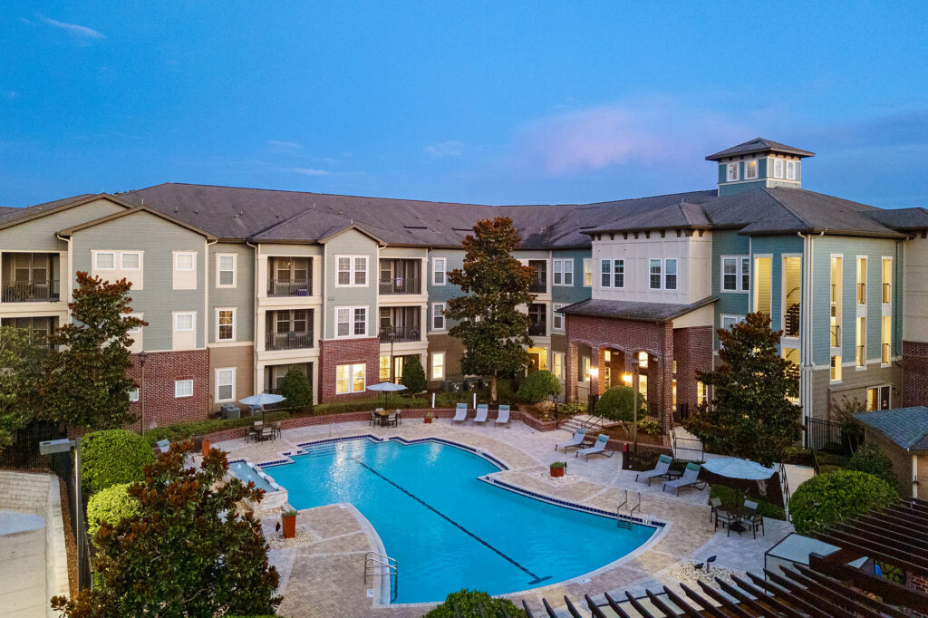 An apartment complex at dusk with a large outdoor swimming pool, lounge chairs, umbrellas, and surrounding greenery. The building has three stories and a tower structure on one end.