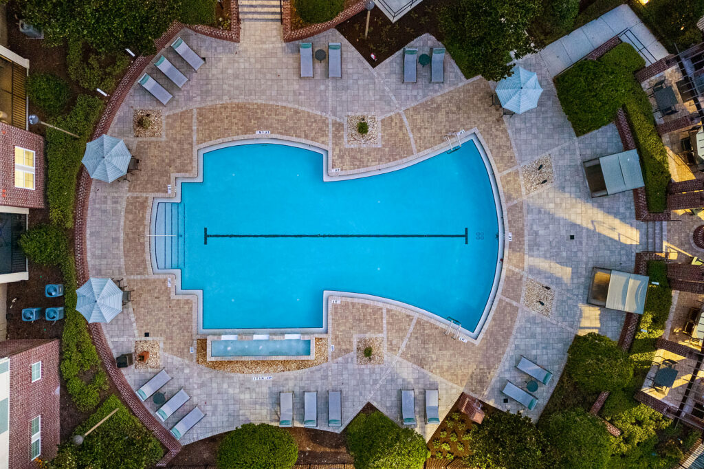 Aerial view of a large, uniquely shaped swimming pool surrounded by lounge chairs, umbrellas, and greenery in a paved courtyard area.