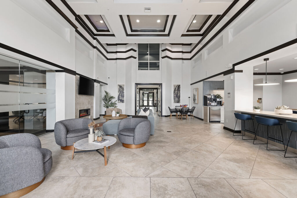 Modern lobby with gray armchairs, coffee tables, tiled floors, a fireplace, bar seating, a coffee station, wall art, and large windows letting in natural light.