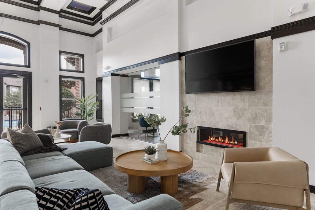Modern living room with a gray sectional sofa, round wooden coffee table, beige armchair, wall-mounted TV, and fireplace. Large windows let in natural light. Neutral tones and minimalist decor create a cozy atmosphere.