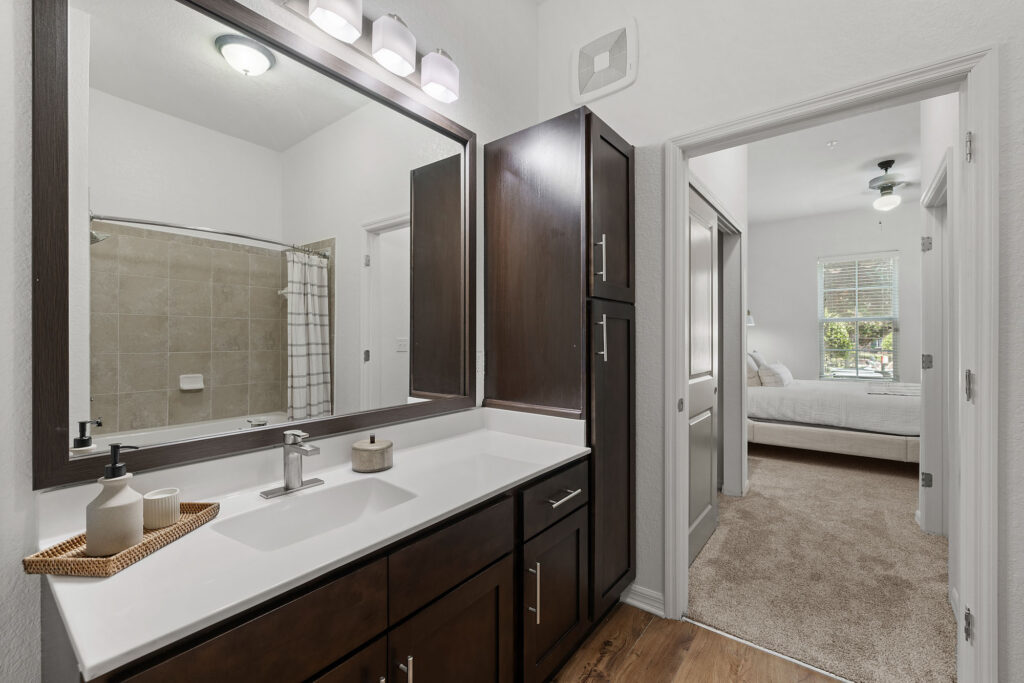 Modern bathroom with a large mirror, double sink vanity, dark cabinets, and a tray with soap dispensers. The room opens to a bedroom with carpeted floors, a bed, and a window letting in natural light.