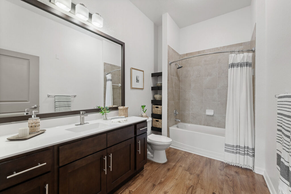Modern bathroom with a large mirror, dark wood cabinets, double sinks, a toilet, and a tub-shower combo with beige tiles and a white shower curtain. Towels and decor add a touch of style to the space.