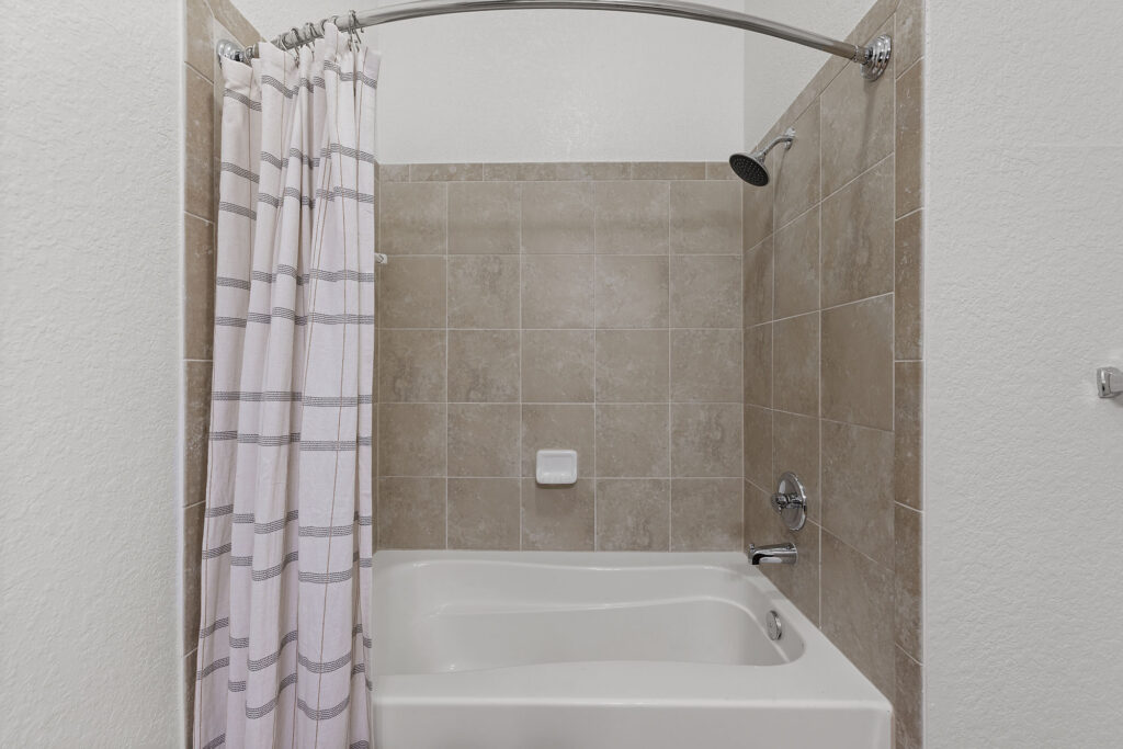 White bathtub with beige tiled walls, a silver showerhead, and a white soap holder. A white and gray striped shower curtain hangs on a curved silver rod. The overall look is clean and simple.