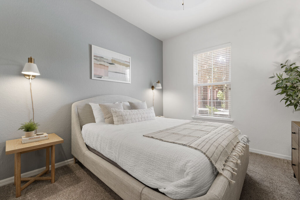 Modern bedroom with a large bed, neutral bedding, a wooden side table with a book and plant, wall lamps, abstract art, window with blinds, and a potted plant in the corner.