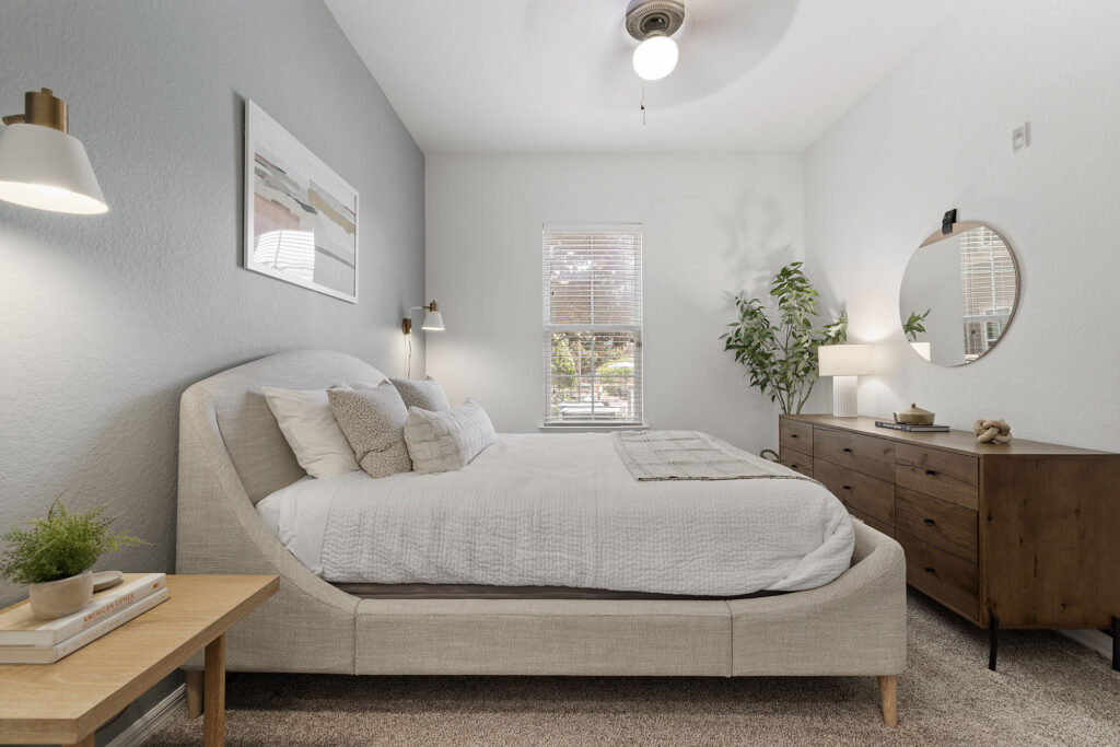A modern bedroom with a large bed, white bedding, wooden nightstand, dresser with round mirror, potted plant, and a window letting in natural light. Neutral colors and minimal decor create a calm, cozy atmosphere.