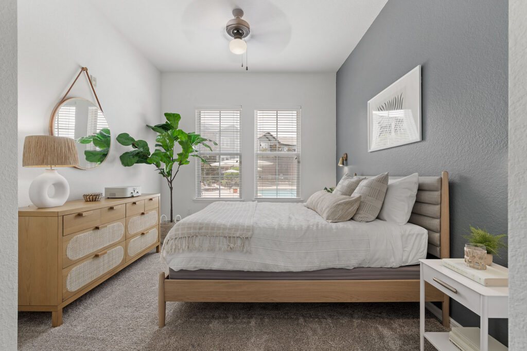 Bright bedroom with a gray accent wall, wooden bed, white bedding, dresser with mirror, potted plant, and large windows letting in natural light, showing a view of a pool outside.