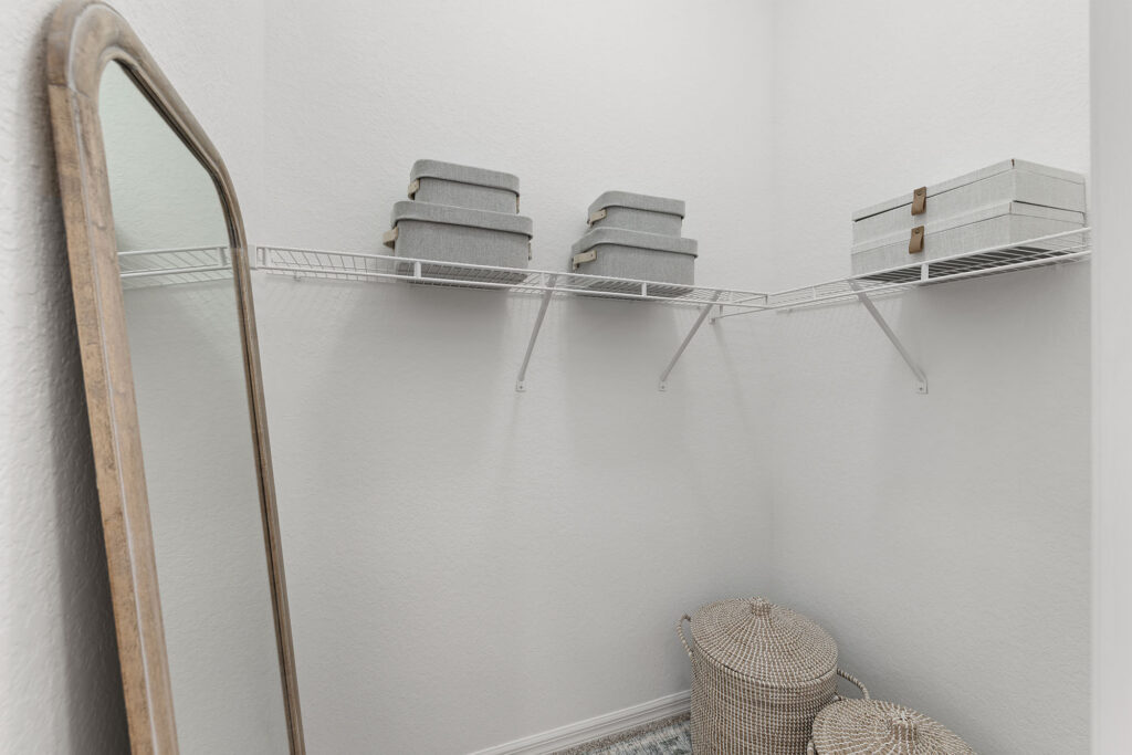 A walk-in closet with white walls, a wire shelf holding gray storage boxes, two woven baskets on the floor, and a tall mirror with a wooden frame leaning against the wall.