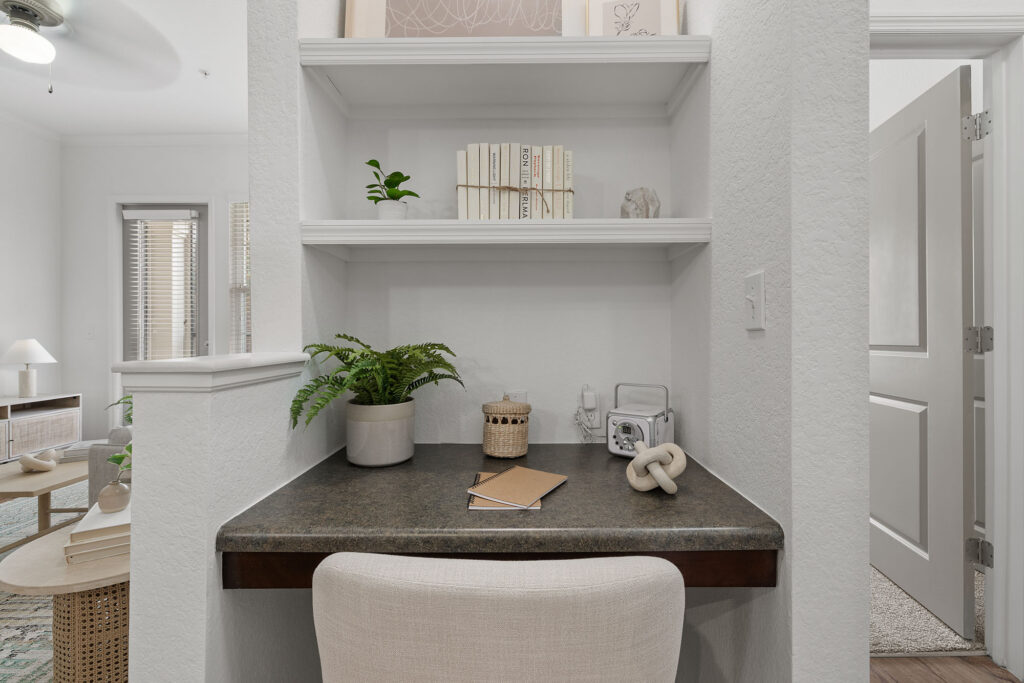 A small built-in desk with a chair, potted plant, decorative items, and notebooks, framed by white shelves holding books and decor; a cozy, modern workspace in a bright room.