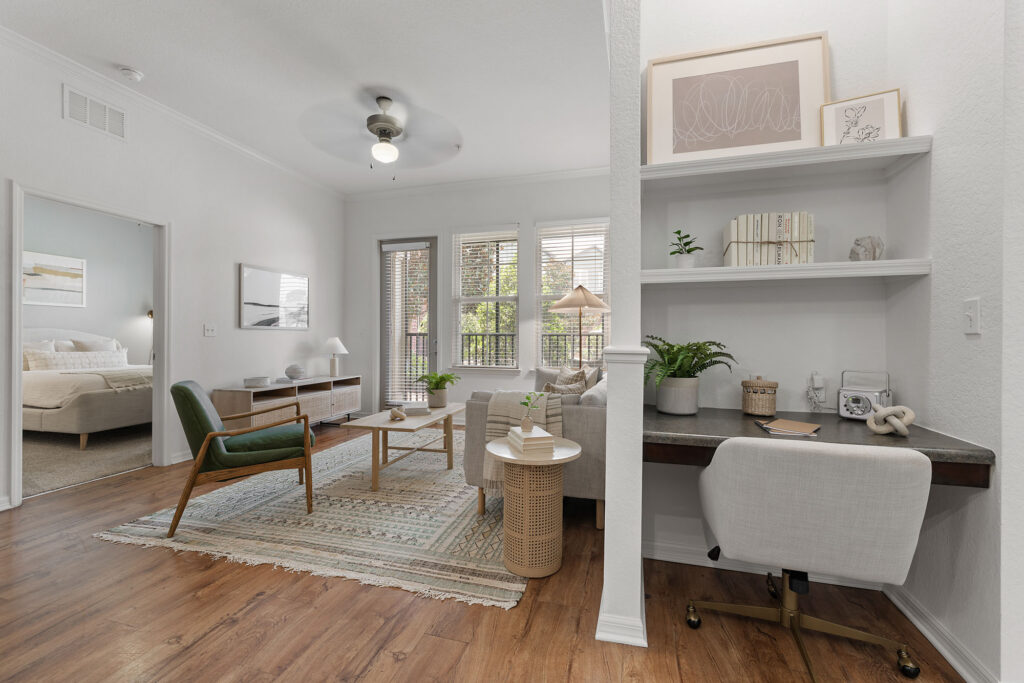 Bright living room with modern decor, wood flooring, a green chair, sofa, coffee table, and rug. A small home office nook with shelves, a desk, and chair sits adjacent. Bedroom visible through open door.