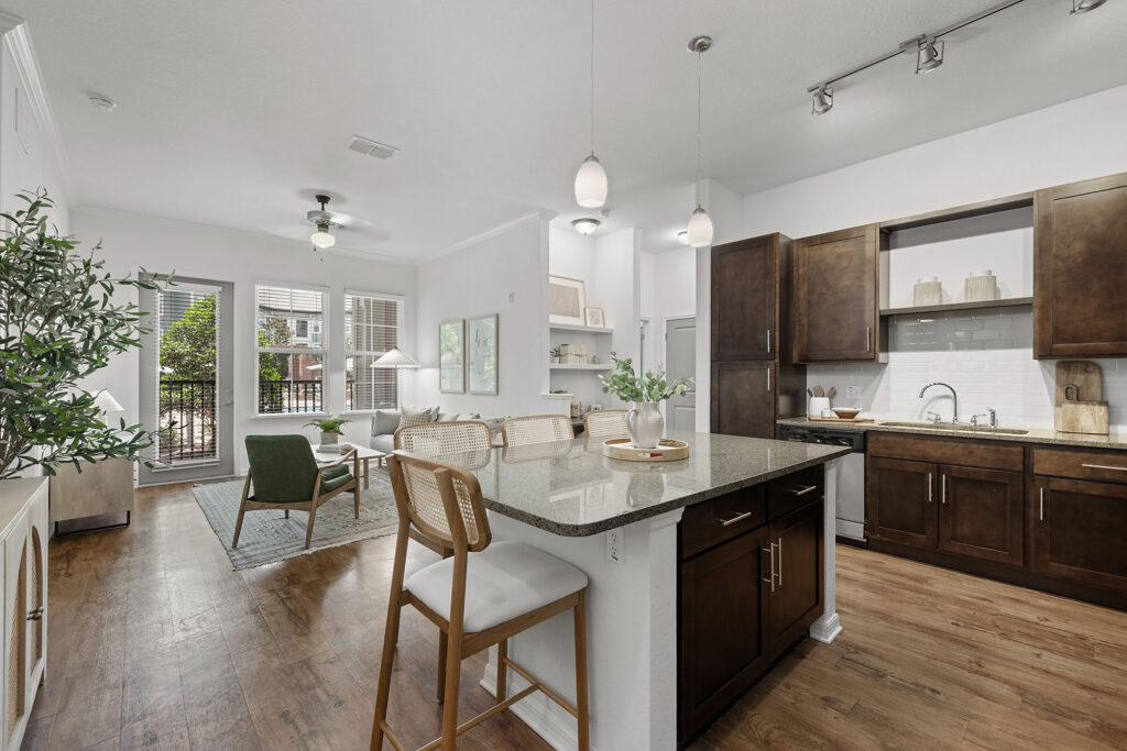 Modern open-concept kitchen and living room with dark wood cabinets, island seating, light wood floors, and large windows letting in natural light. Sliding doors open to a patio with greenery outside.