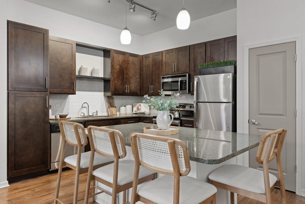 Modern kitchen with dark wood cabinets, stainless steel appliances, an island with four wooden chairs, overhead lighting, and minimalist decor, including a vase with greenery on the counter.