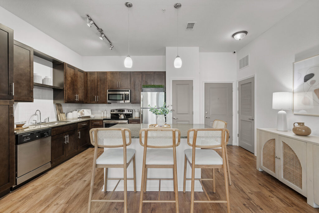 Modern kitchen with dark wood cabinets, stainless steel appliances, a white island with four barstools, wood flooring, and minimalist decor. Two pendant lights hang above the island.