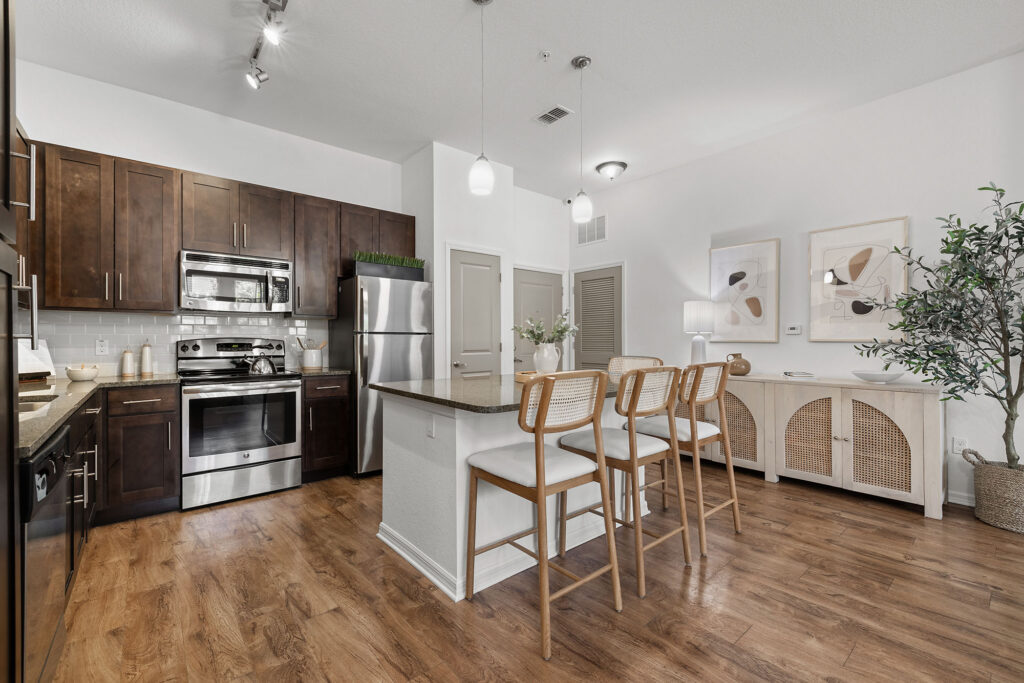 Modern kitchen with dark wood cabinets, stainless steel appliances, a small island with three wooden barstools, hardwood floors, and a cozy dining area with wall art, a sideboard, and a potted plant.