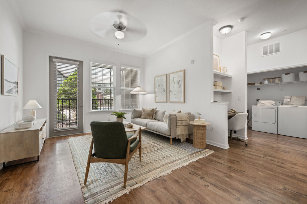 Modern living room with a rug, green chair, sofa, and lamp. Large windows let in light. In the background, theres a small office nook and laundry area with a washer and dryer. Hardwood floors throughout.