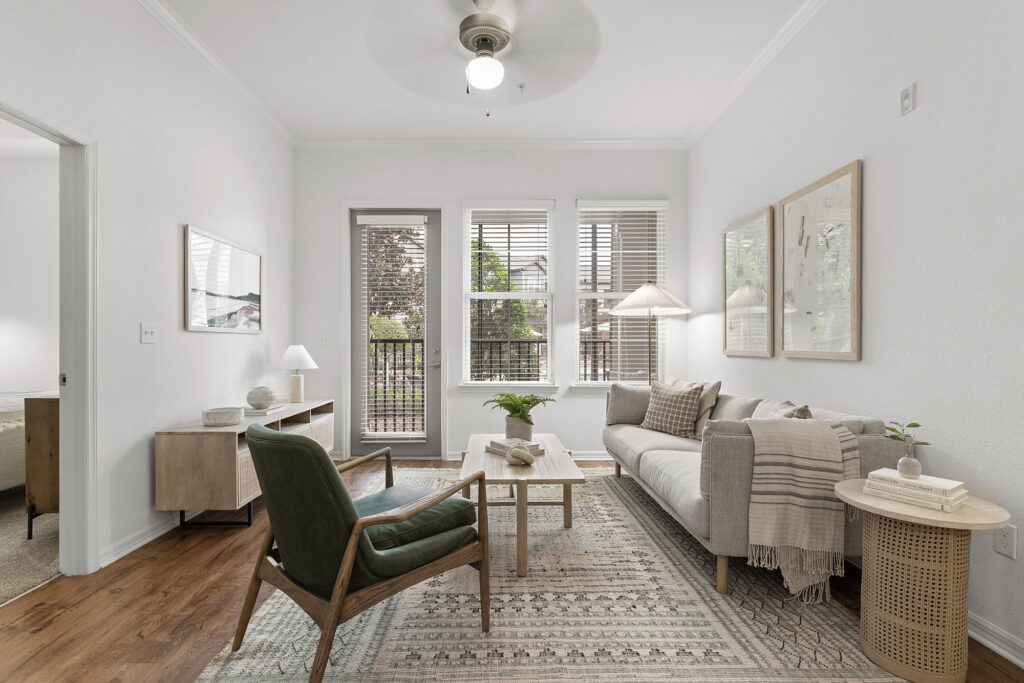 Bright living room with a sofa, green armchair, wooden coffee table, floor lamp, side tables, and artwork on white walls. Large windows let in natural light and offer a view of greenery outside.