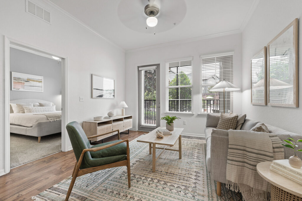 Bright living room with a green armchair, gray sofa, wooden coffee table, and large windows; bedroom visible through open doorway. Neutral decor and a ceiling fan complete the cozy, modern space.