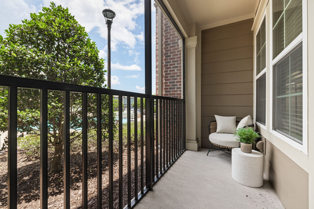 A cozy screened-in patio with a black railing, potted plant, and cushioned chair. A leafy shrub and lamppost are outside, and the sky is partly cloudy. Large windows line the building wall.